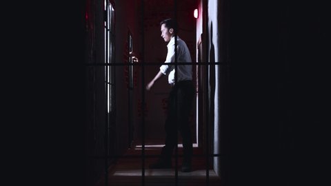 Horror acting - a dangerous male warden walks by the cells in the prison holding a metal pipe and beats the hands of the prisoner on the bars with a pipe