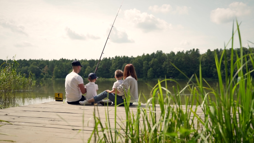 Young happy family with little boy fishing a camping site. Parents with kids fishing together sitting on a wooden pier by pond spending weekend holiday outdoors. Back Rear view. catching the fish