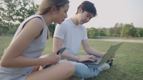 A young happy teenagers sitting with working on computer notebook in green park, a cheerful people having a good time on remote working outside, people laughing and talking together, wireless devices