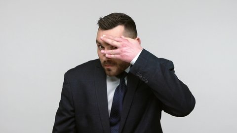 Bearded businessman in black official style suit covering eyes with palm, peeking through fingers, looking with suspicion, shy to watch forbidden event. Indoor studio shot isolated on gray background.