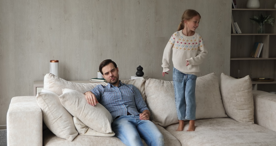Young tired father sit on sofa at home feels exhausted by noisy little daughter makes noise jumping near irritated annoyed dad. Parent and difficult hyperactive misbehaved child need attention concept Royalty-Free Stock Footage #1078337864