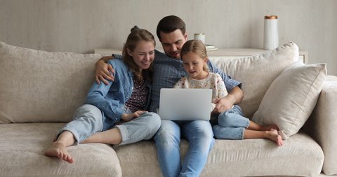 Happy couple and little 8s daughter relax on sofa at home using laptop, having fun on internet, discuss new video game enjoy leisure with on-line fun. Young generation and modern wireless tech concept