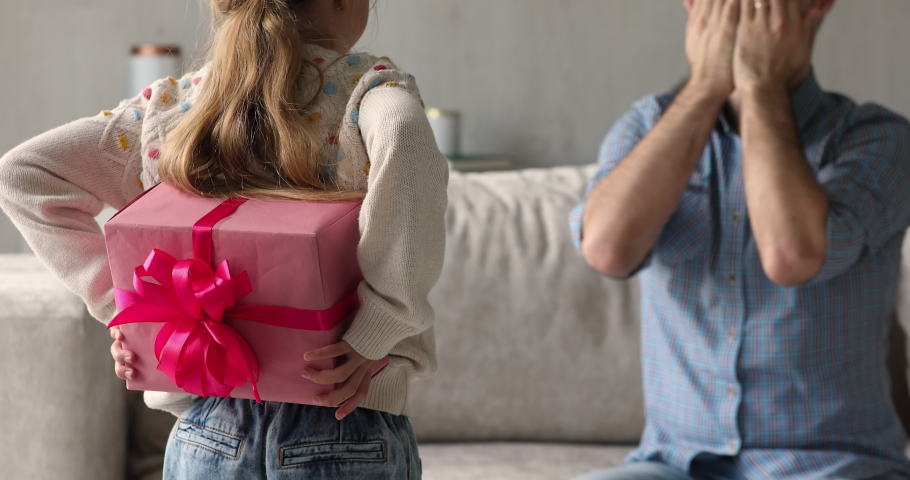 Little loving daughter hides box, standing in front of daddy sits on sofa, cover eyes anticipate moment of presenting gift. Happy Father day celebration and congratulation, family life events concept Royalty-Free Stock Footage #1078337936