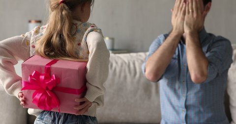 Little loving daughter hides box, standing in front of daddy sits on sofa, cover eyes anticipate moment of presenting gift. Happy Father day celebration and congratulation, family life events concept