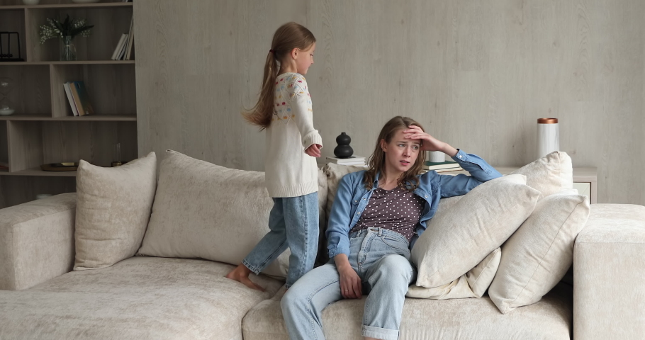 Exhausted woman sit on sofa feels irritated by loud hyperactive little daughter jumps makes noise nearby. Single divorced helpless mom and her hyperactive kid, upbringing problem, bad behavior concept Royalty-Free Stock Footage #1078338011