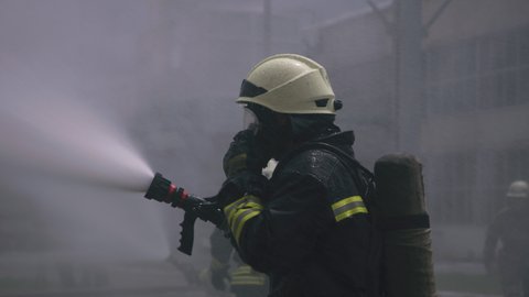 Anonymous firefighter adjusting respirator while pouring water from hose during training in fire station yard