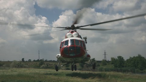 Tracking shot of red and white emergency service helicopter taking off from grassy ground against cloudy blue sky on sunny day in countryside