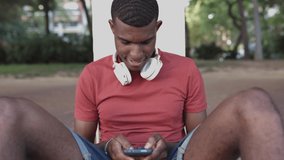 Hispanic young man with headphones using mobile phone while sitting on the ground in city street - Social media, new trends technology and millennial generation concept