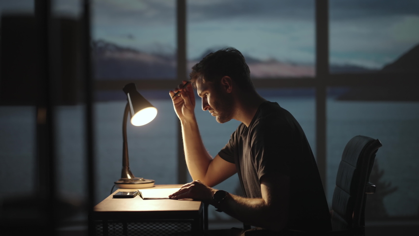 The silhouette of a thoughtful man sitting at a table against the background of a window with oceans and the sea. A restless man sitting at a table with a desk lamp | Shutterstock HD Video #1078347893
