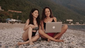 Two women sitting on the beach and talking on video conference with friends. People using laptop to communicate with family. Social distancing during pandemic. Coronavirus concept.