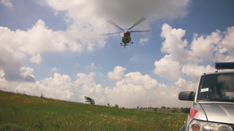 Modern helicopter flying over grassy ground near car against cloudy blue sky during rescue mission in countryside