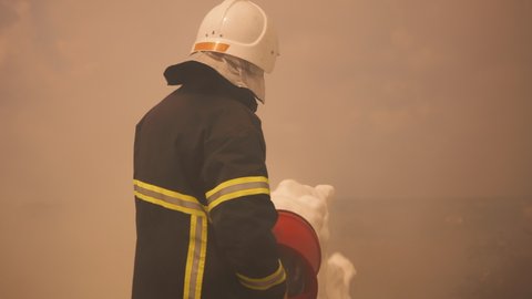 Firefighter in protective uniform and helmet pouring foam from hose to ground while standing in thick fume and extinguishing fire