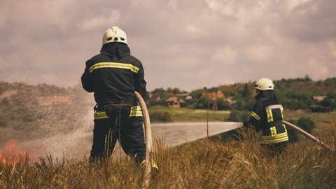 Unrecognizable firefighters with hoses walking in field and pouring water on burning dry grass during rescue mission in countryside