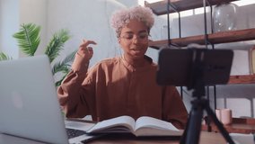 Young friendly African American woman recording video blog. Biracial female sitting at home office with laptop and book, recording video on mobile phone camera that stands on desk on mini tripod.