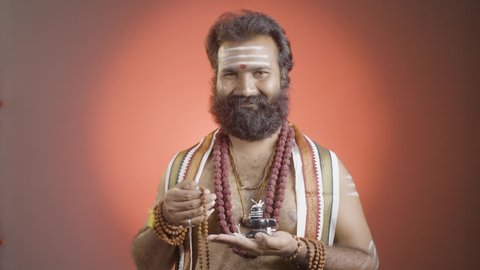Indian priest or holy guru meditating or chanting using rudrakshi japa mala with shiva linga in hand on studio background