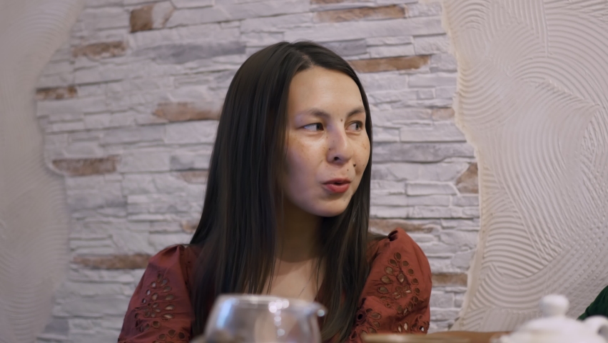 A beautiful native American girl of appearance is talking at the table. The girl has many moles on her face, slanted eyes and dark hair.