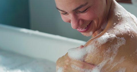 Cinematic close up of young woman washing her shoulder while relaxing in foamy bubble filled perfumed bathtub at home. Concept of purity, personal hygiene, spa resort, comfort, skincare, healthcare.