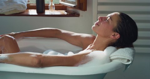 Cinematic shot of young woman is relaxing with pleasure in foamy bubble filled perfumed bathtub at home. Concept of purity, personal hygiene, spa resort, comfort, skincare, healthcare.