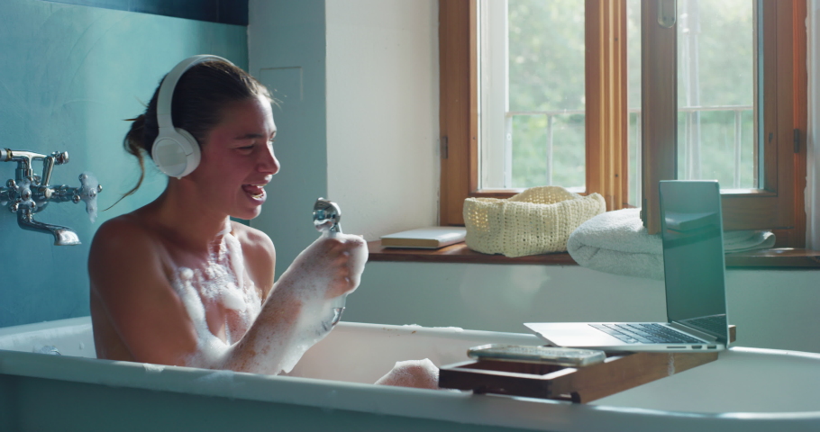 Portrait of Woman With Headphones, Smartphone and Laptop, Sitting in a Bathtub, Listening to Music, Singing Along. Female in Foamy Bubbles Pretending to be a Performing Singer. Shower Karaoke Moment 