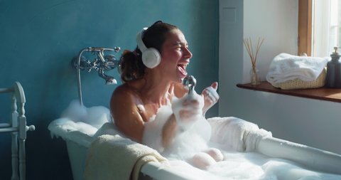 Authentic shot of carefree woman having fun to listen music and sing with shower handle while relaxing in foamy bubble filled bathtub at home. Concept: leisure, freedom, happiness, comfort, skincare.