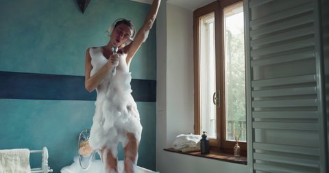 Authentic shot of carefree woman having fun to listen music, sing with shower handle and dance crazy in foamy bubble filled bathtub at home. Concept: leisure, freedom, happiness, comfort, skincare.
