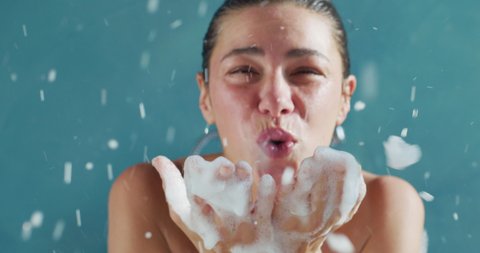 Authentic shot of happy young woman blowing foamy soap bubbles in camera while relaxing in bathtub at home. Concept of purity, happiness, personal hygiene, spa resort, comfort, skincare, healthcare.