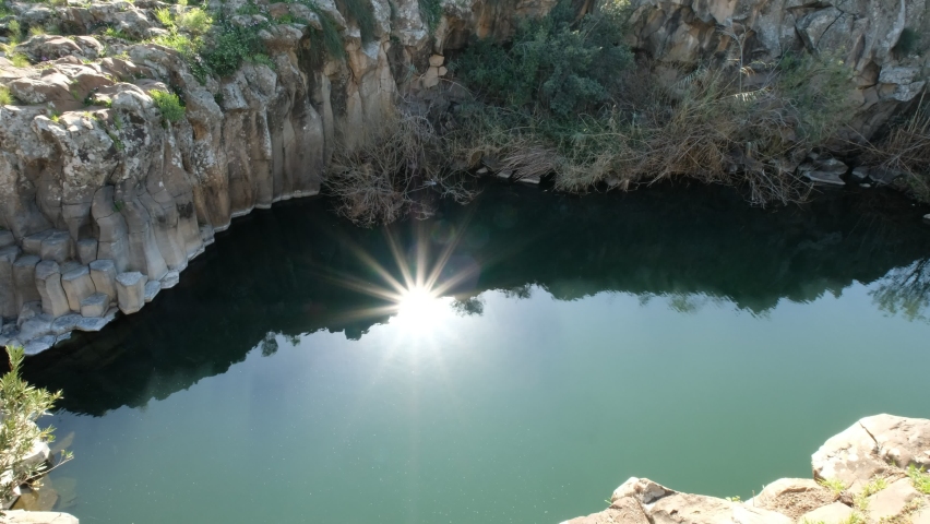A natural, wild water pool in a basalt surroundings with hexagon shapes walls. a 4K handheld shot of the serene environment, with fantastic sun glitter upon the water. Golan heights, Israel.