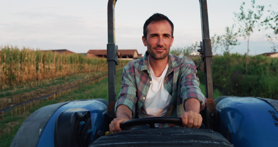 Medium Shot of a Handsome Middle-Aged Man Driving a Tractor on a Road by a Green Field of Corn. Professional Male Farmer Starting his Work Tasks in the Early Morning. He is Checking his Crops Royalty-Free Stock Footage #1078375712