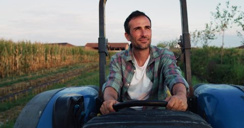 Cinematic shot of male farmer is driving tractor and smiling satisfied with his work on countryside road of farmland corn fields. Concept: agriculture, industry, cultivation, bio and eco farming.