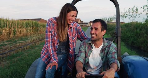 Cinematic shot of happy couple of farmers driving tractor and smiling satisfied with work on countryside road of farmland corn fields. Concept: agriculture, industry, cultivation, bio and eco farming.