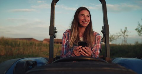 Cinematic shot of female farmer using smartphone in tractor and smiling satisfied on countryside farmland corn fields. Concept:technology, connection, machinery, agriculture, industry, modern farming