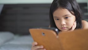 closeup asian child or kid girl lying on bed watching to playing tablet or learn from home and study online on computer or people e-learning communication by smartphone pad in bedroom with copy space