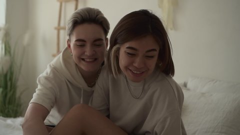 Lesbian couple sit relax in bed with dog. Positive smiling women with cute jack russell domestic pet in morning. Female in cozy modern room interior. Concept romantic, same sex relationship