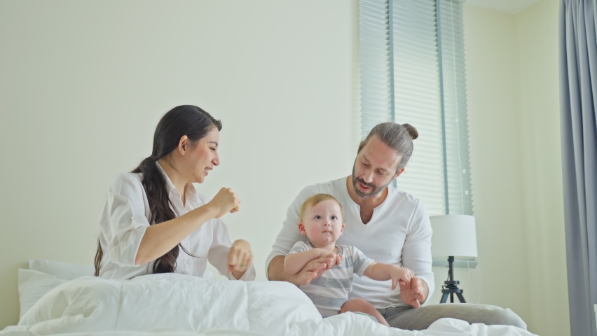 Caucasian loving parents play with baby boy child on bed in bedroom. Happy family, attractive beautiful young couple dad and mom dance with cute infant toddler son enjoy morning activity after wake up
