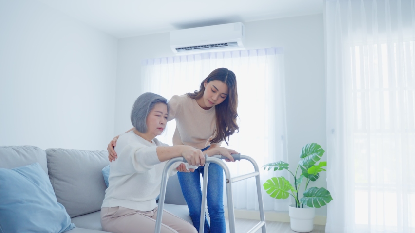 Asian young daughter support old disabled woman walk with walker at home. Beautiful girl help and take care of senior elderly mature handicap mom patient doing physical therapy in living room in house | Shutterstock HD Video #1078386857