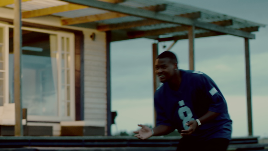 HANDHELD African American father and son playing football together on a lawn in front of their house in the evening. Shot with 2x anamorphic lens