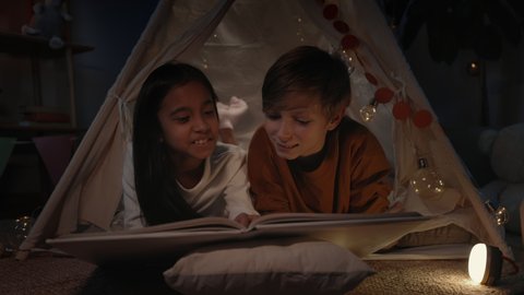 Teens in decorative makeshift tent turning page while reading book and smiling in evening. Cheerful boy and girl lying on floor while spending free time. Concept of leisure and carelessness