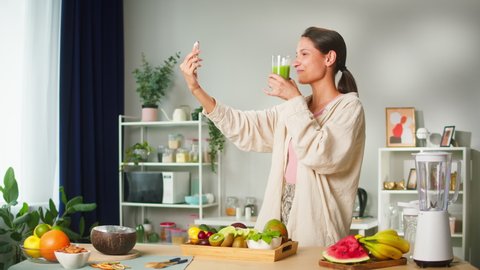 Young smiling woman speaking on video call and drinking juice, food blogger recording video vlog on smartphone how to cook smoothie. Healthy sweet detox cocktail. 