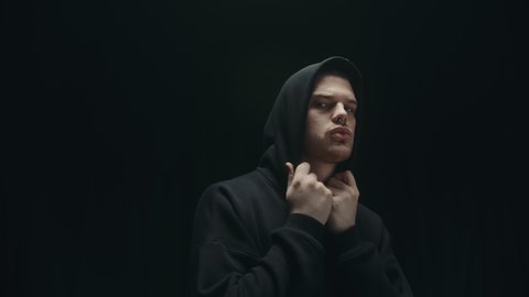 Fashion portrait of brutal man putting on his hood, strong muscle guy with nose piercing in dark hoodie and modern trendy hairstyle. Model posing, fashion studio concept. 