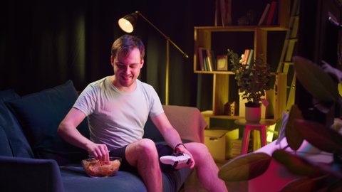 Young man gamer playing video game on tv and eating snack chips in evening. Guy winner using modern console, sitting on sofa in living room. Cybersport, gaming at home. Professional player