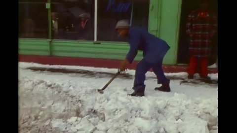 CIRCA 1977 - A man shovels outside his shop in Buffalo, New York, while professionals use snow plows on the street.