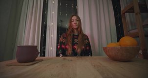 Young emotional woman in Christmas pullover talks by videocall. Emotional girl with active facial expressions. Christmas lights decorated in dining room at home.