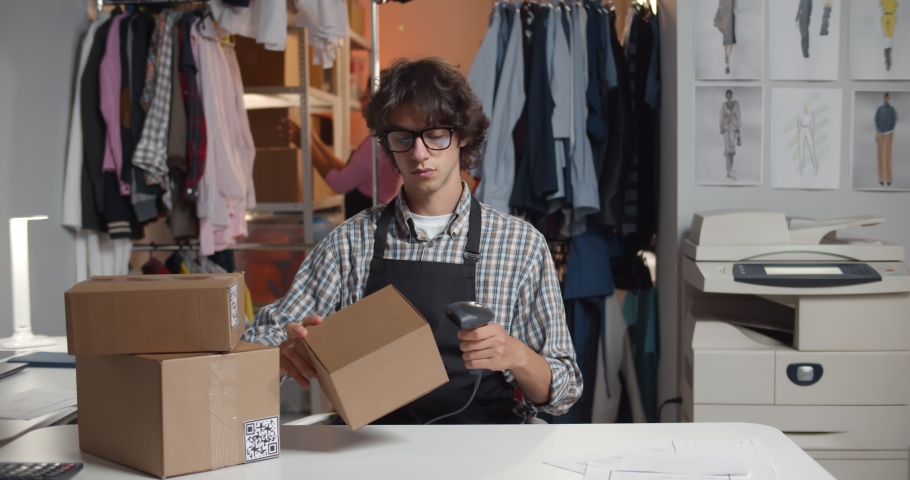 Male online clothes store worker scanning parcel bar code packing order. Portrait of young man sitting at desk in modern atelier workshop preparing online retail shop order Royalty-Free Stock Footage #1078399799