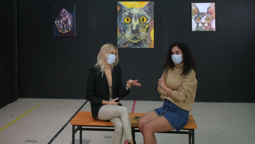 Two friends having a talk at the art exhibition. One woman giving her arguments and trying to prove her point of view. Visitors in medical masks, COVID-19 restrictions