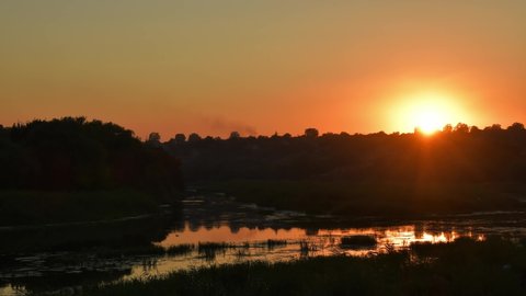 Sundown time lapse. Orange color sunset sky over Southern Bug river shore with sun reflection in rippled water overgrown by reeds and bulrush plants. Tranquil landscape Bug Guard National Park Ukraine
