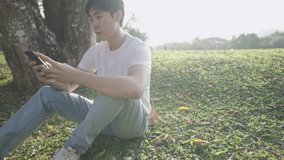 Young asian male using smartphone sitting under big tree inside the park, hot sunny day, staring at phone, portable device, mobile phone and modern technology, application subscription plan, solitude