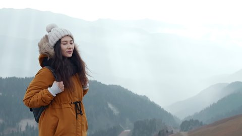 Happy asian woman tourist enjoys breathing fresh air high in cloudy mountains at cold weather. Young hiker at amazing hazy background of mountain landscape in smoke. Concept of active lifestyle