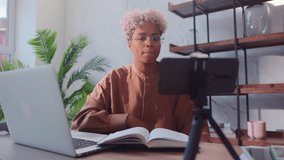 Happy african american woman turns on cell phone camera to record video. Beautiful young woman sitting with book and laptop and going to record useful video for her blog using phone on tripod.
