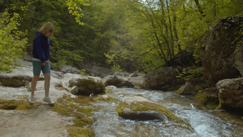 Female hiker walking on rocks at river. Smiling woman with backpack hiking by mountain river. Happy girl travel in green landscape in mountains