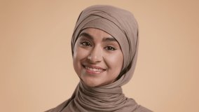Portrait Of Happy Middle-Eastern Millennial Lady Wearing Hijab Posing Standing Over Beige Studio Background, Smiling To Camera. Beauty Of Muslim Females Concept. Slow Motion